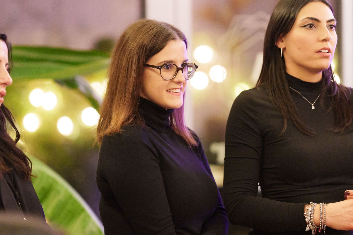 Three women in black outfits at an indoor event, smiling, with bokeh lights in the background.