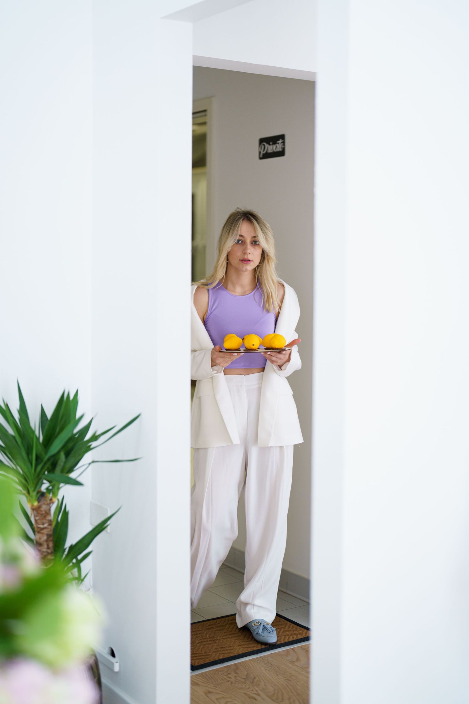Blonde woman in white wide-leg pants and lavender crop top holding a plate of oranges in a bright doorway.