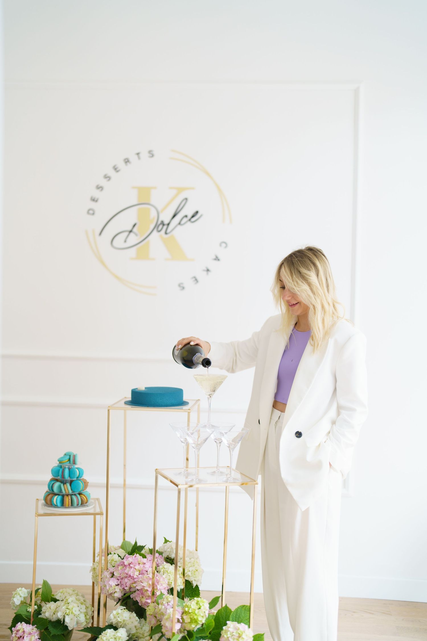 Woman in white suit pouring champagne at K Dolce Desserts and Cakes bakery with blue cake and macarons.