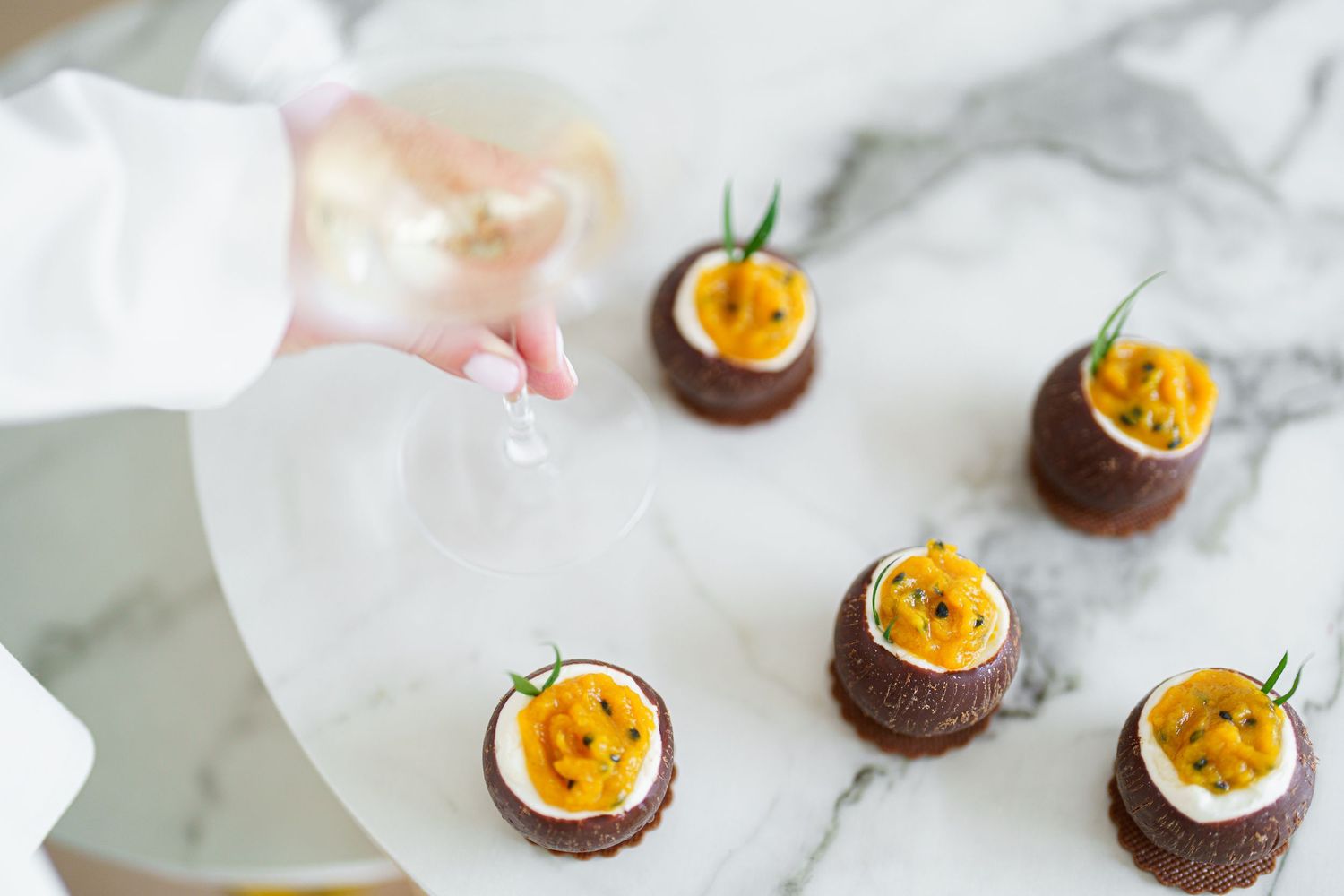 Hand placing garnish on elegant chocolate appetizers topped with vibrant yellow passion fruit on a marble serving plate.
