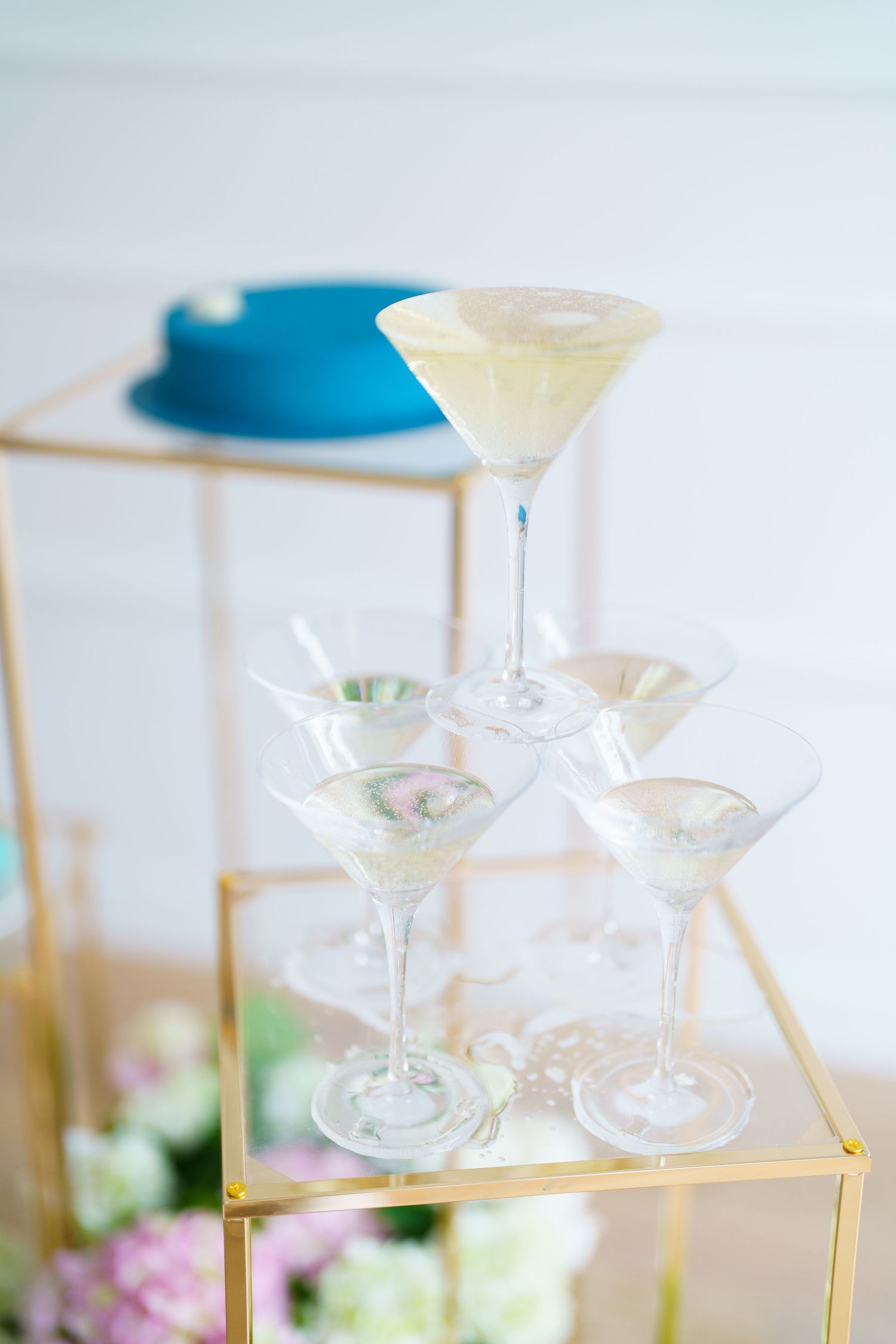 Elegant champagne tower with martini glasses on a gold stand, blue cake in background, floral decorations at a wedding reception.