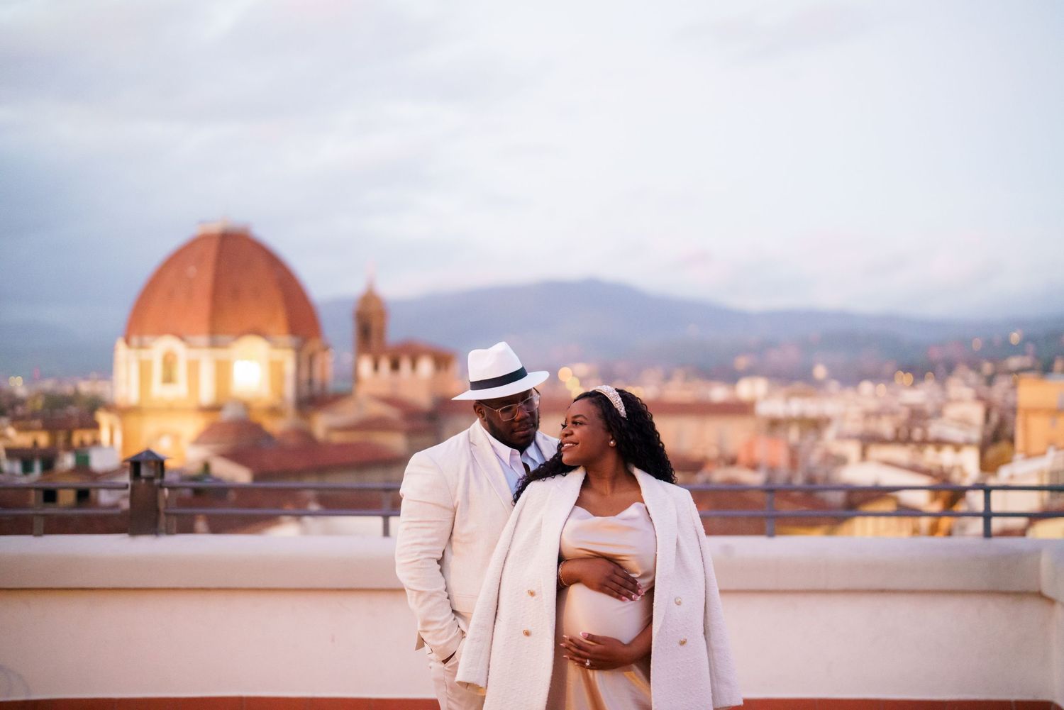 Couple in elegant white attire sharing a romantic moment on a rooftop in Florence, Italy, with the Duomo at dusk.