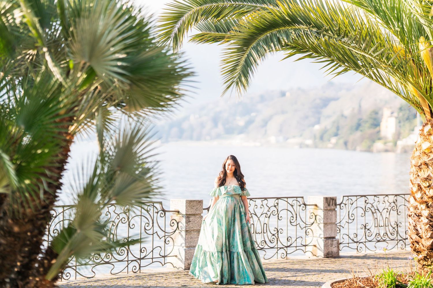 Woman in elegant teal gown poses on ornate lakeside terrace framed by lush palm trees with scenic mountain backdrop.