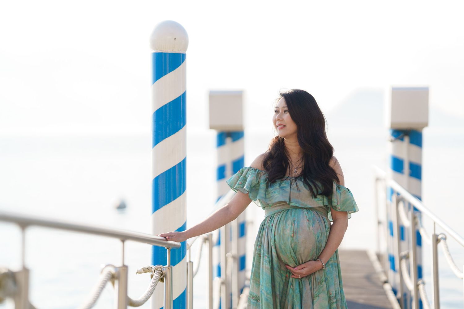 Pregnant woman in teal dress on seaside pier with blue and white striped poles, smiling serenely at ocean view.
