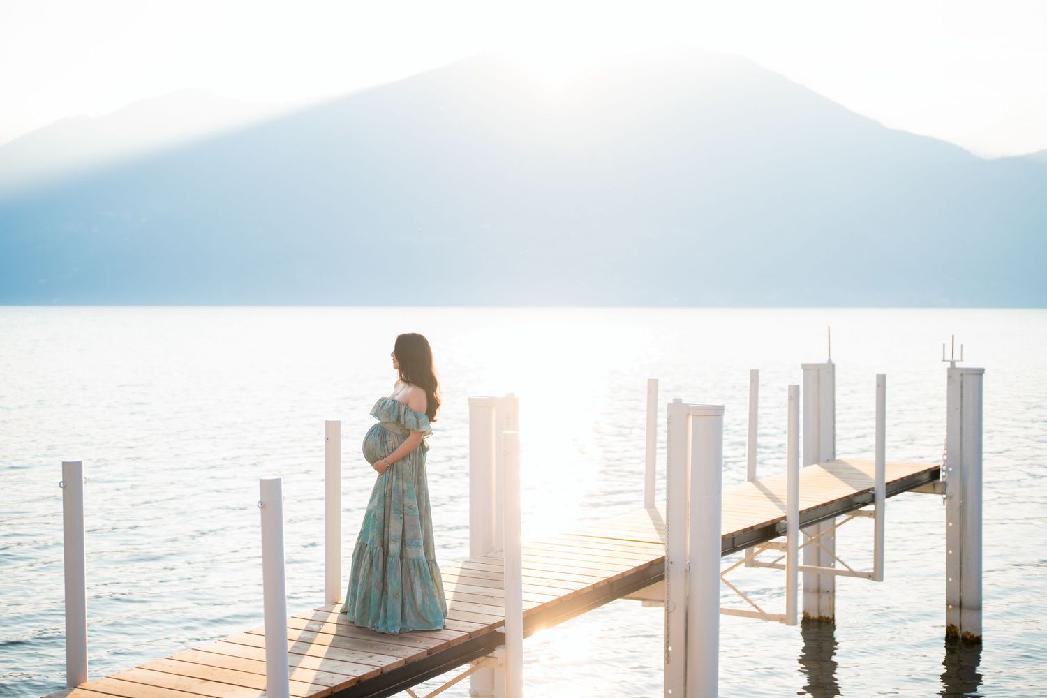 Pregnant woman in blue floral dress standing on wooden dock overlooking calm lake with mountain backdrop.