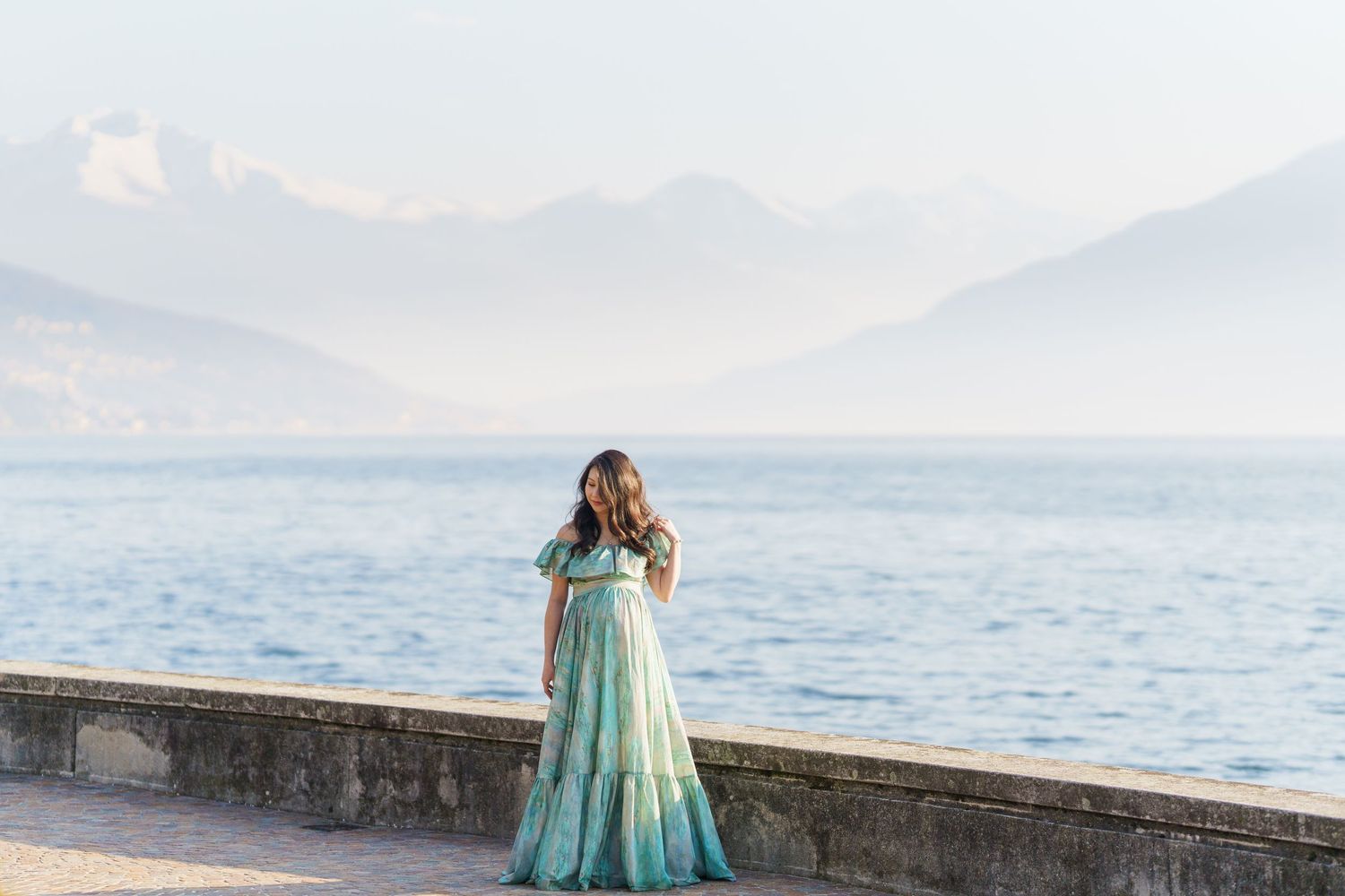 Woman in teal gown stands on lakeside promenade with misty mountains and serene blue water in background.