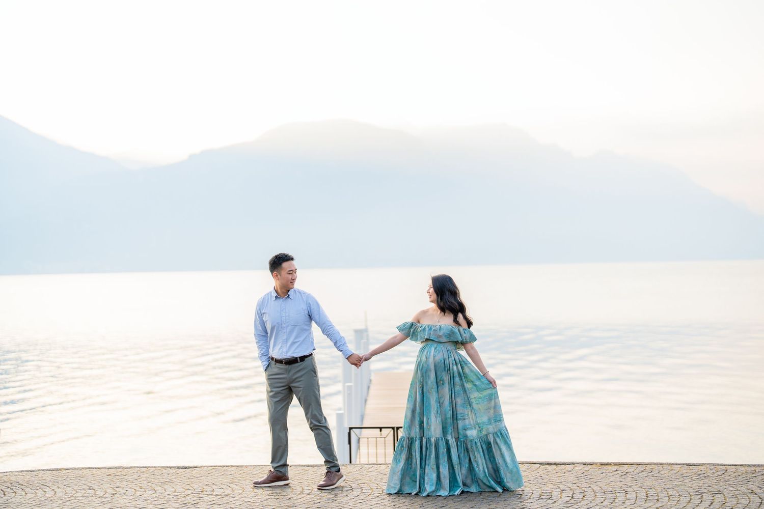 Couple holding hands by a serene lake with misty mountains, woman in teal gown, man in blue shirt.