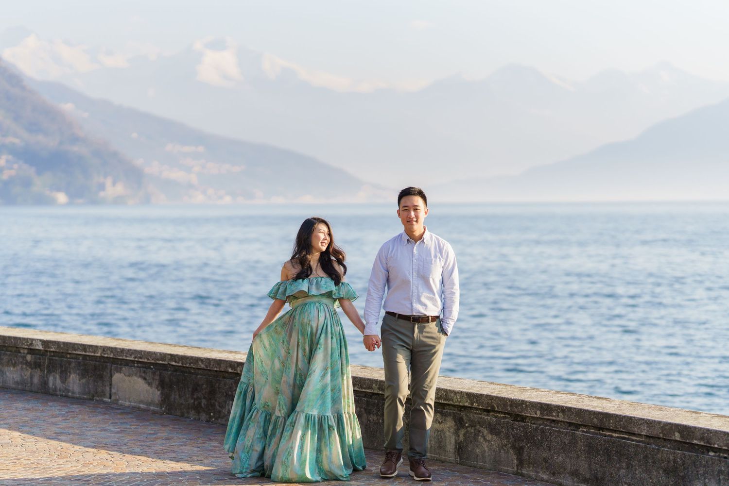 Couple holding hands by Lake Como with mountains, woman in teal maternity gown, man in white shirt and gray pants.