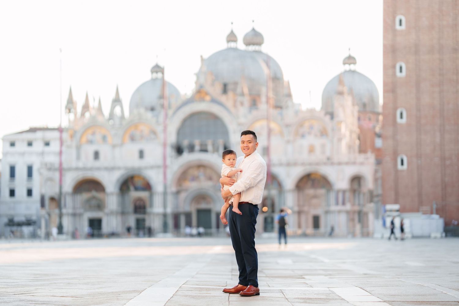 Father holding baby in St. Mark's Square, Venice, with the iconic Basilica di San Marco in the background.