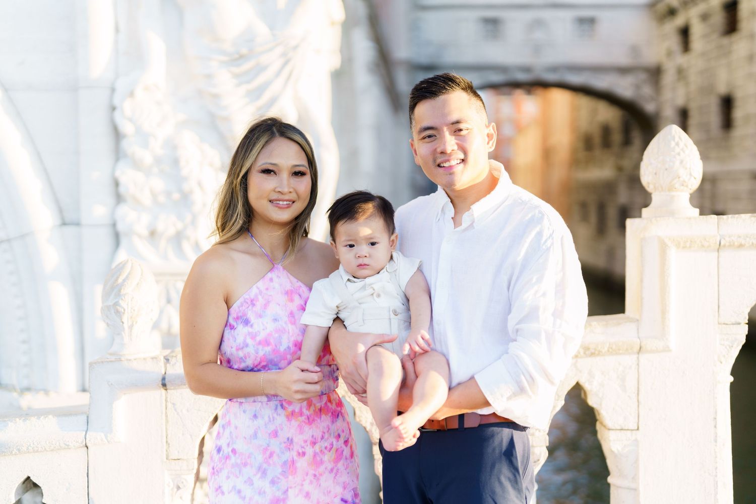 A smiling Asian family with a baby poses outdoors near white ornate architecture in a bright, elegant setting.