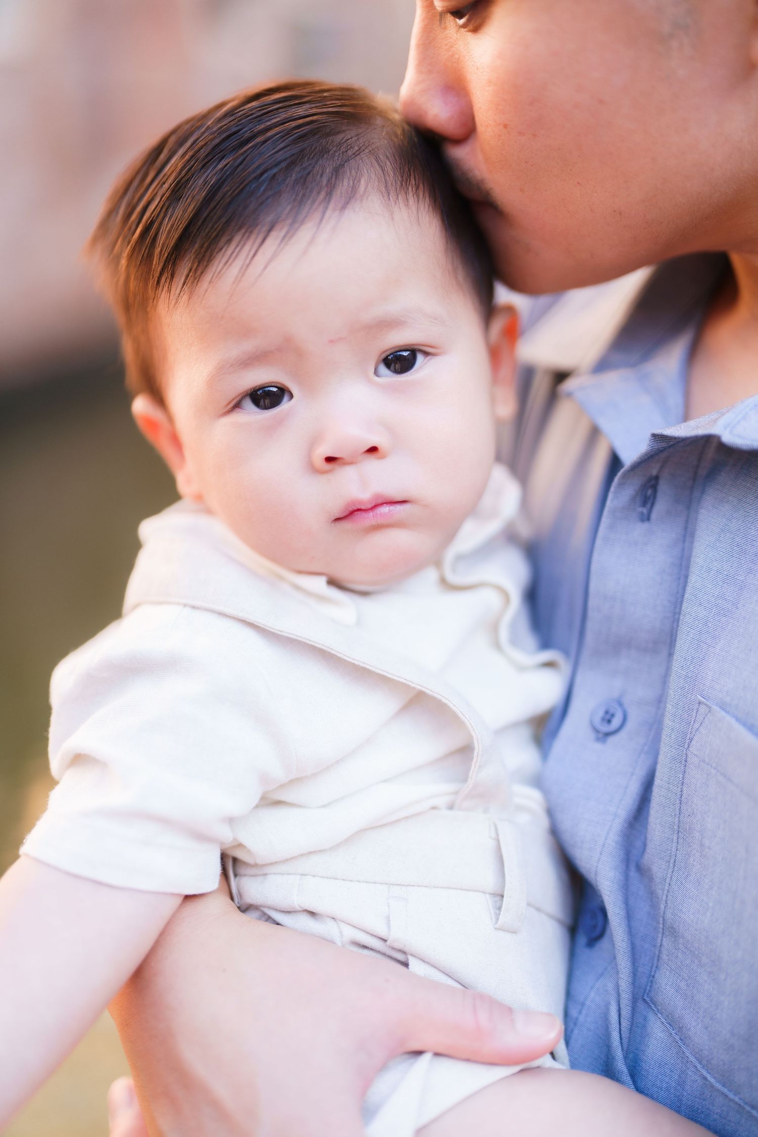 A tender moment as a parent kisses their baby, who wears a white outfit and gazes forward with a calm expression.