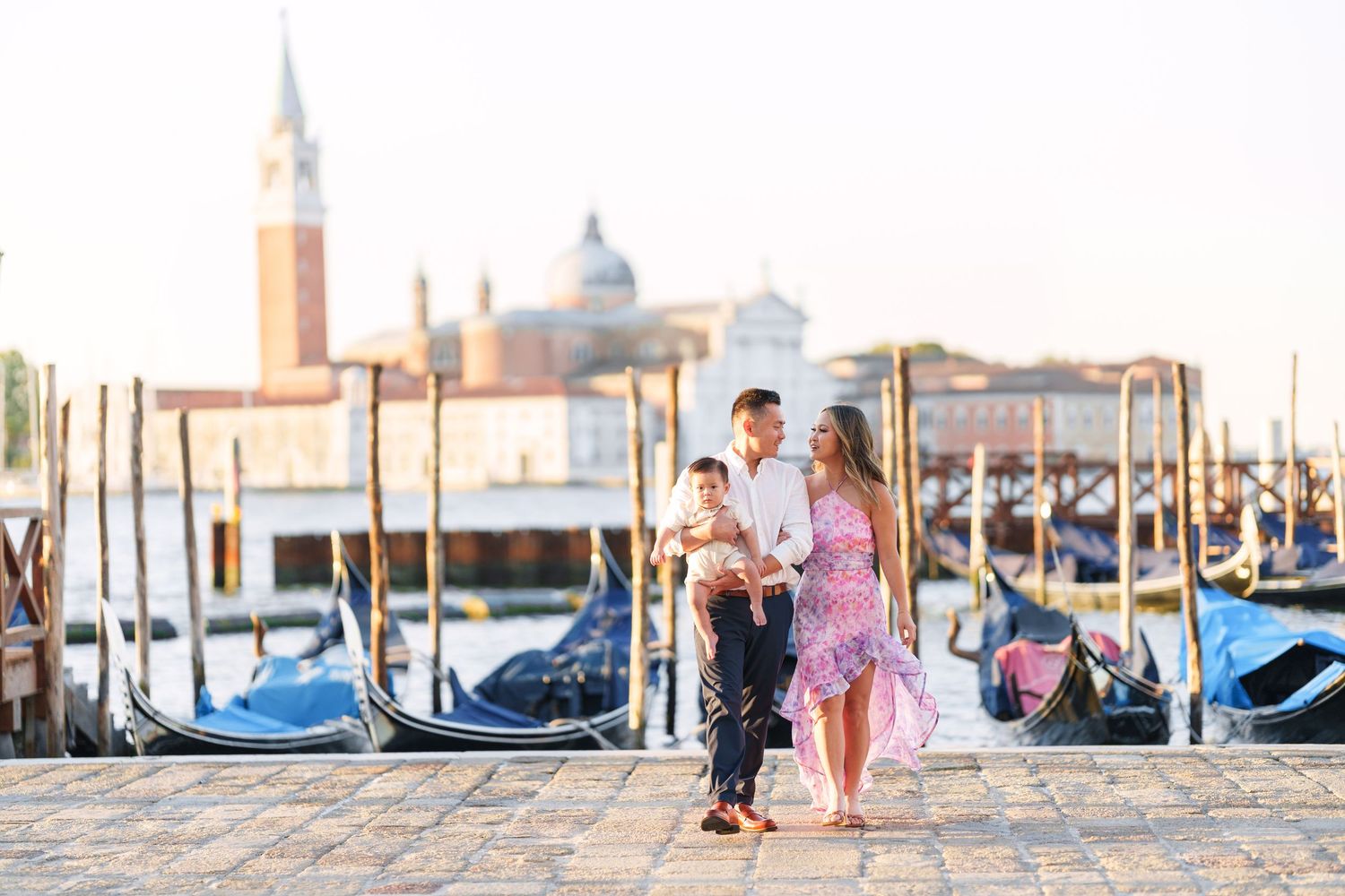 Family of three posing by Venice gondolas with San Giorgio Maggiore church in golden morning light.