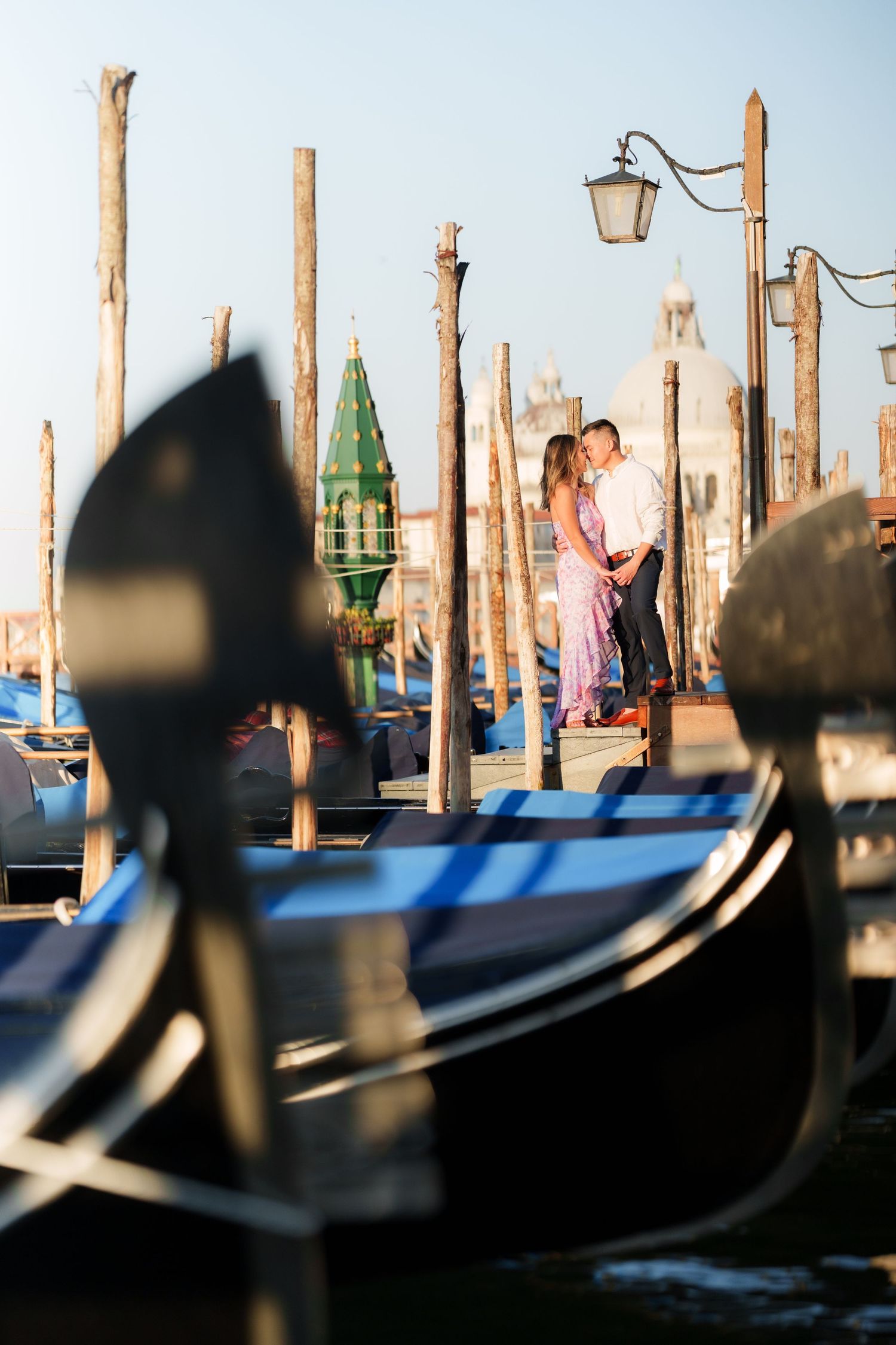Romantic couple embracing among Venice gondolas and wooden poles at golden hour near St. Mark's Square.