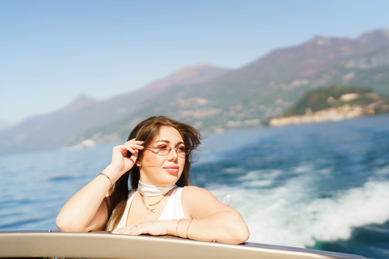 Woman in white top adjusting sunglasses on a boat with mountain lake scenery in the background.
