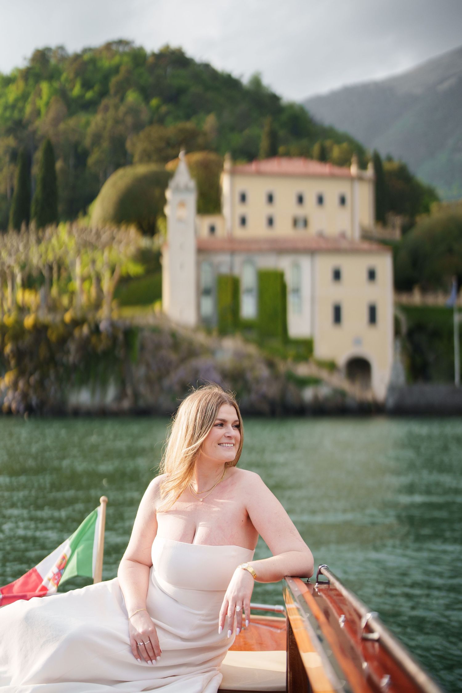Bride in strapless gown sits on wooden boat at Lake Como, Italy, with Villa del Balbianello in background.