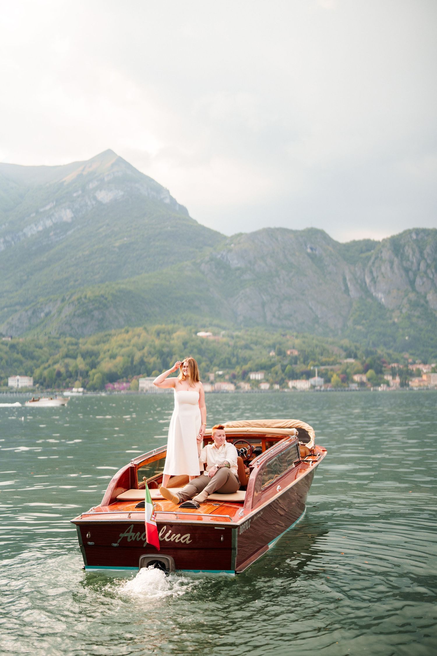 Couple on a classic wooden boat on Lake Como, Italy, with lush green mountains in the background.