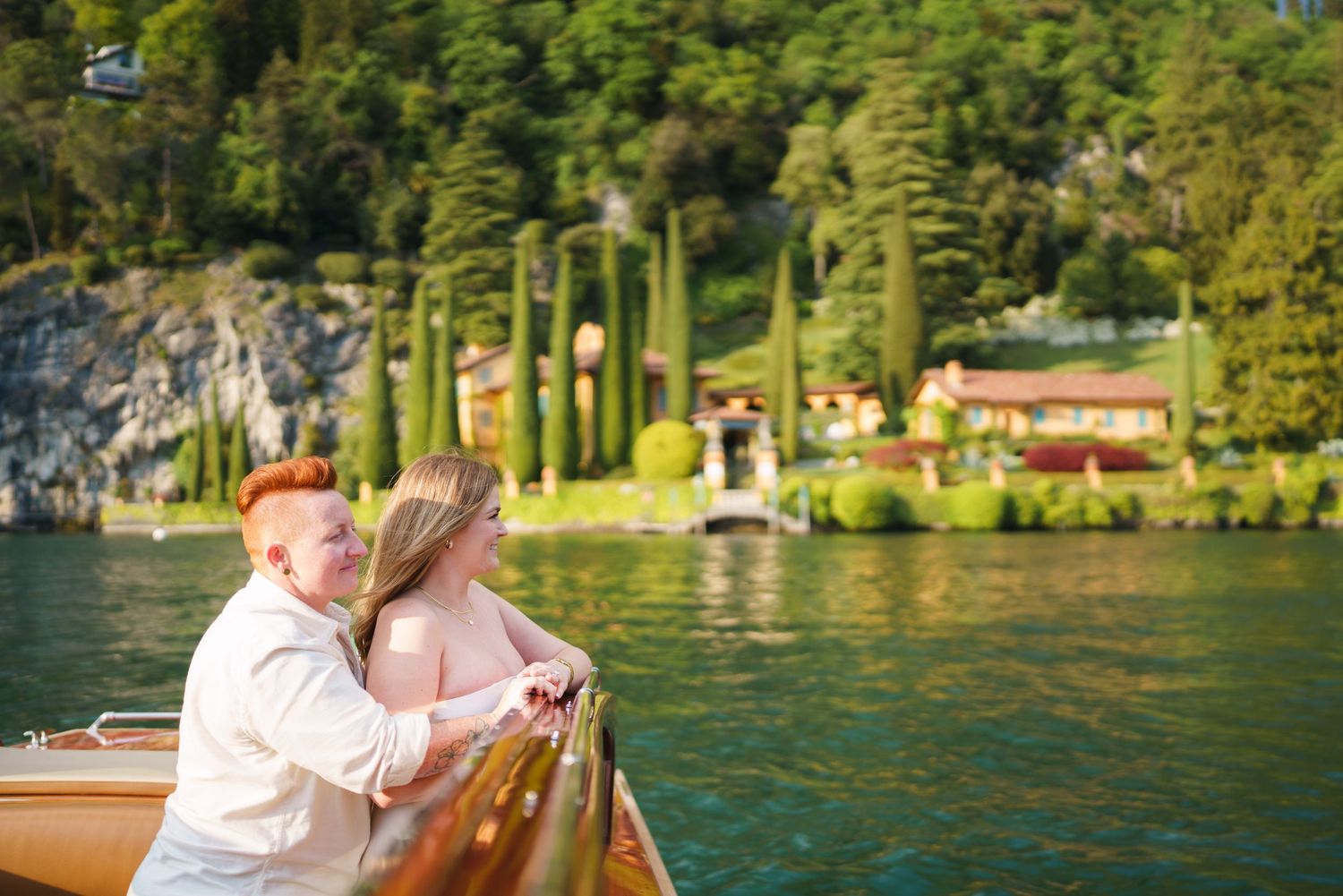 Couple sharing a romantic moment on a wooden boat on turquoise Lake Como, Italy, with lush greenery and villas behind.