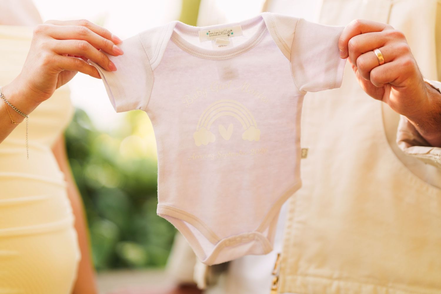 Couple holding a white baby onesie with rainbow print, announcing pregnancy outdoors.