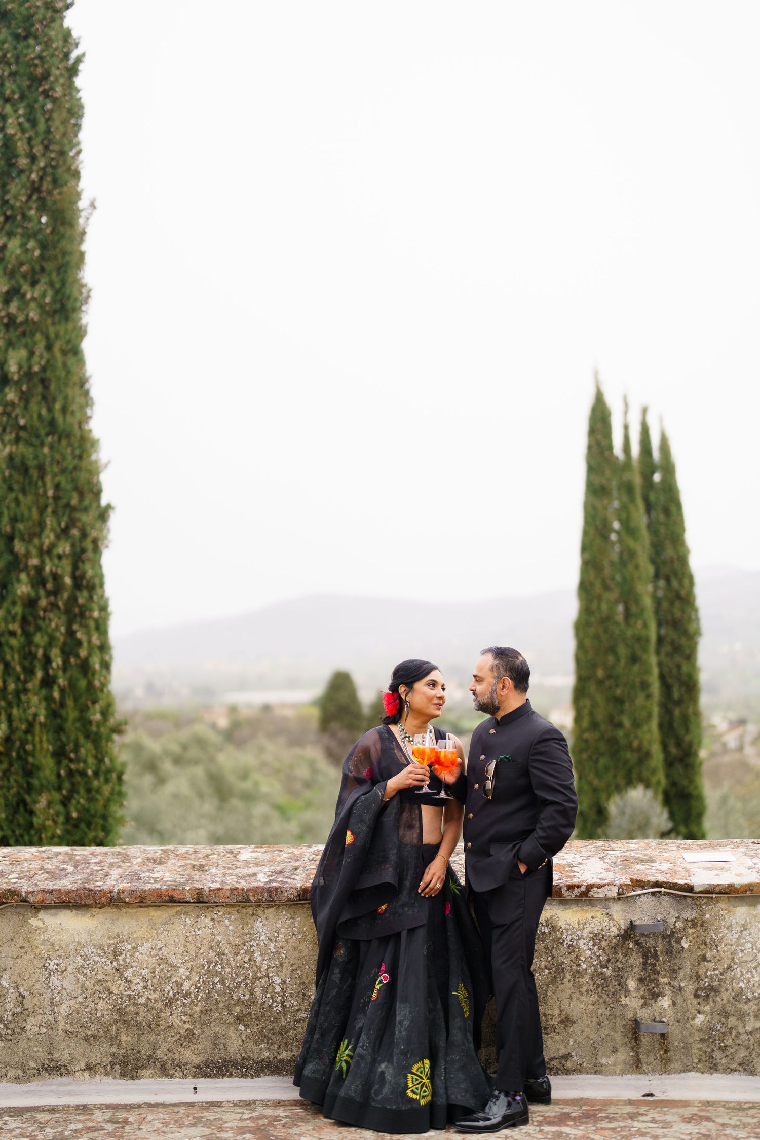Couple sharing a romantic moment against a stone wall with cypress trees and mountains in the background.