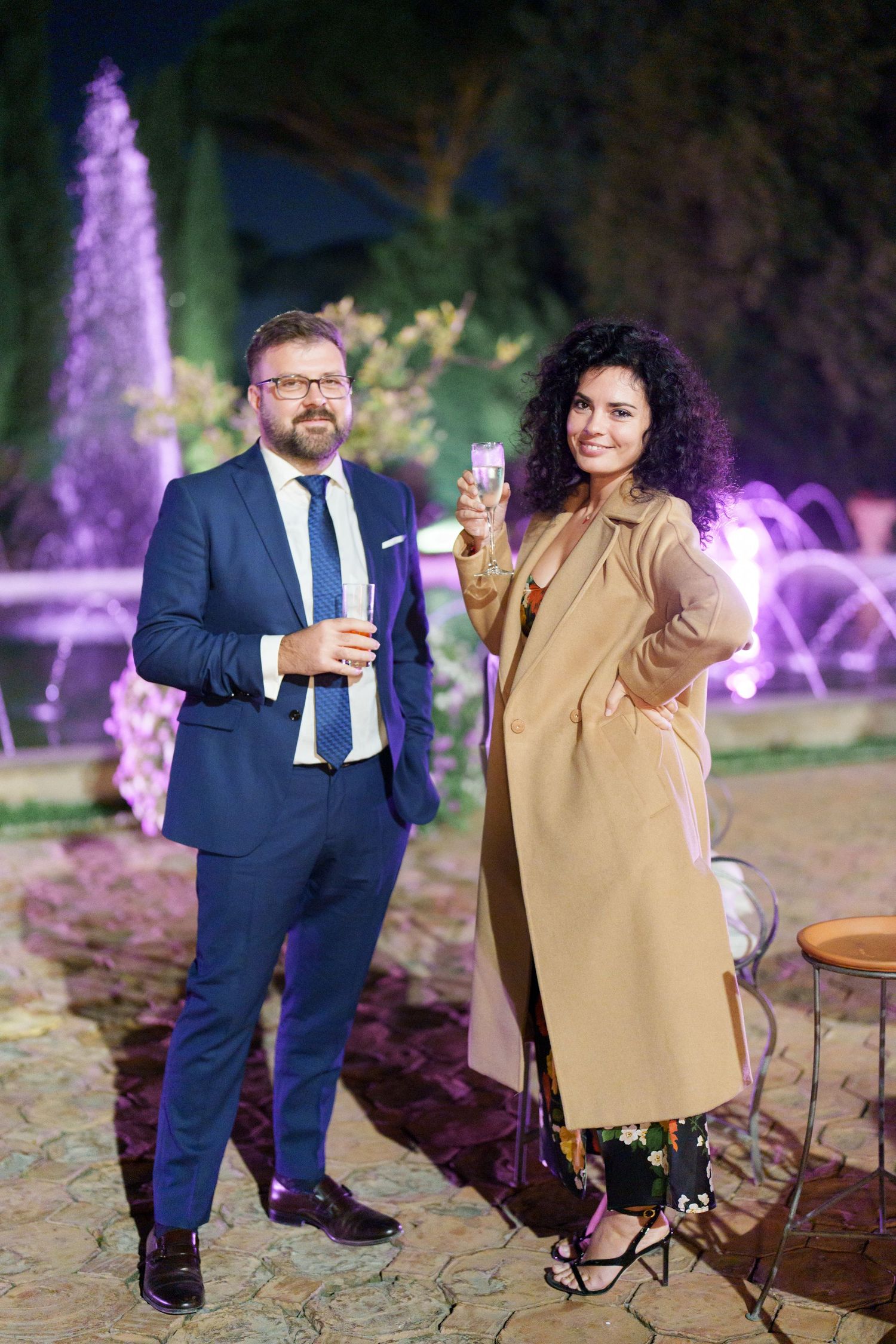 Two guests enjoying drinks at an outdoor evening event with purple fountain lights in the background.