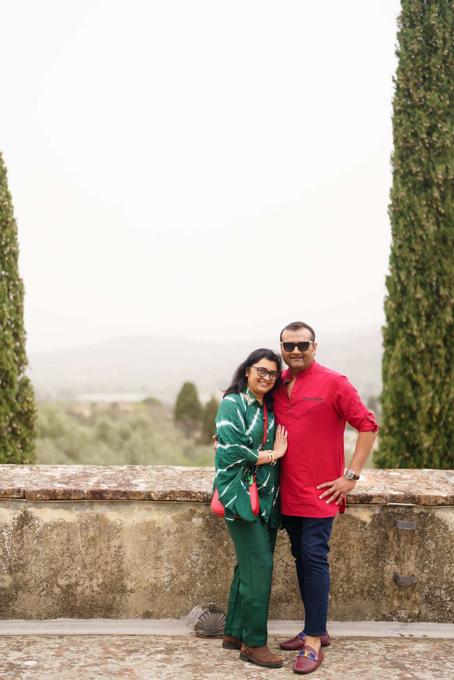 A couple poses casually against a stone wall with Italian cypress trees in the background.