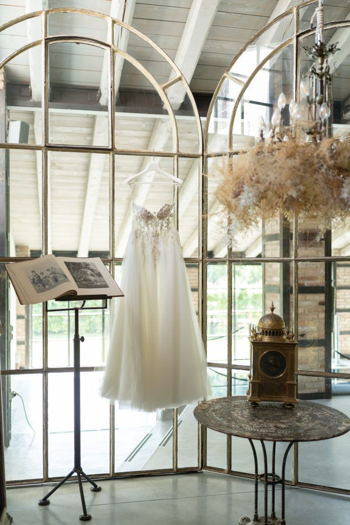 White wedding dress hanging in sunlit room with vintage decor and dried flowers.