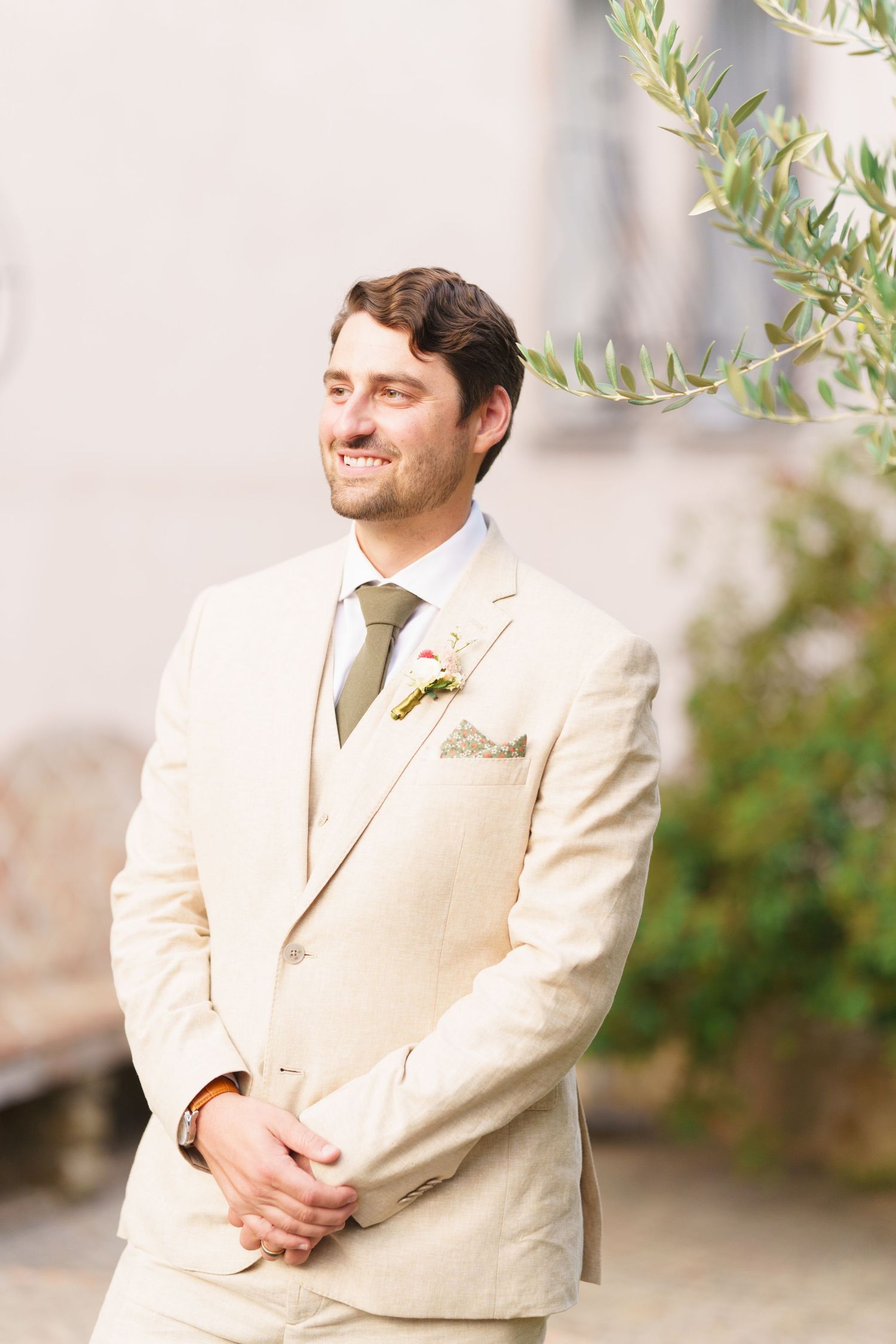 Groom in cream colored suit standing against stone wall backdrop at outdoor venue.