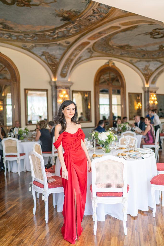 Red formal dress stands out against white dining chairs in luxurious ballroom with ornate ceiling details.