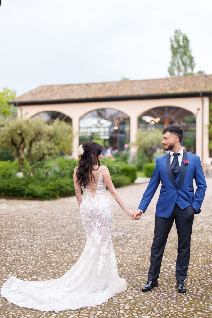 A couple in wedding attire poses together on a cobblestone path near a Mediterranean-style building.