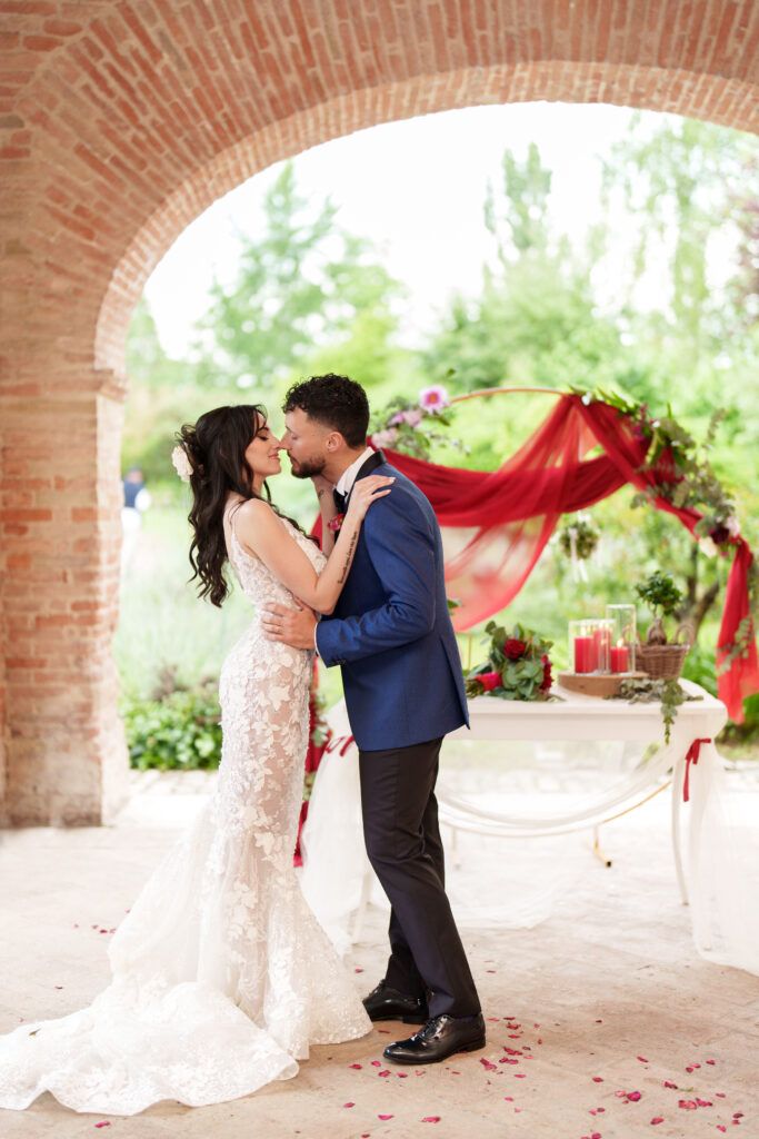 Romantic wedding ceremony under brick archways decorated with flowing red fabric and rose petals.