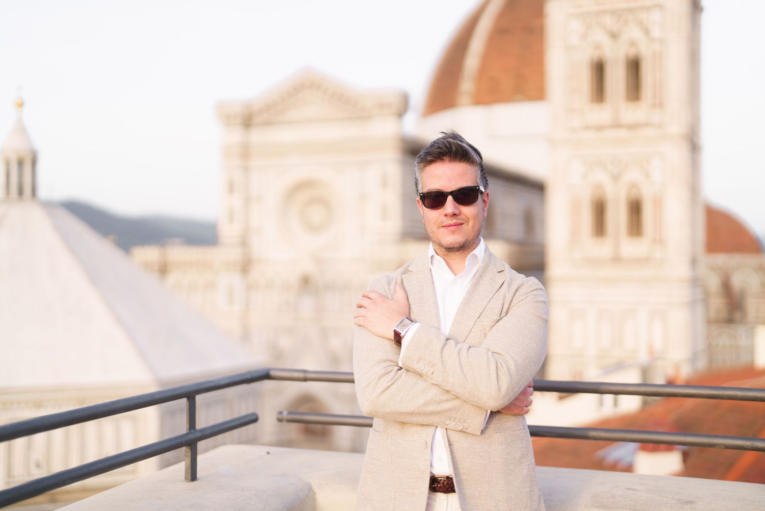 Person in beige suit stands against ornate European architecture background with church spires and domes.