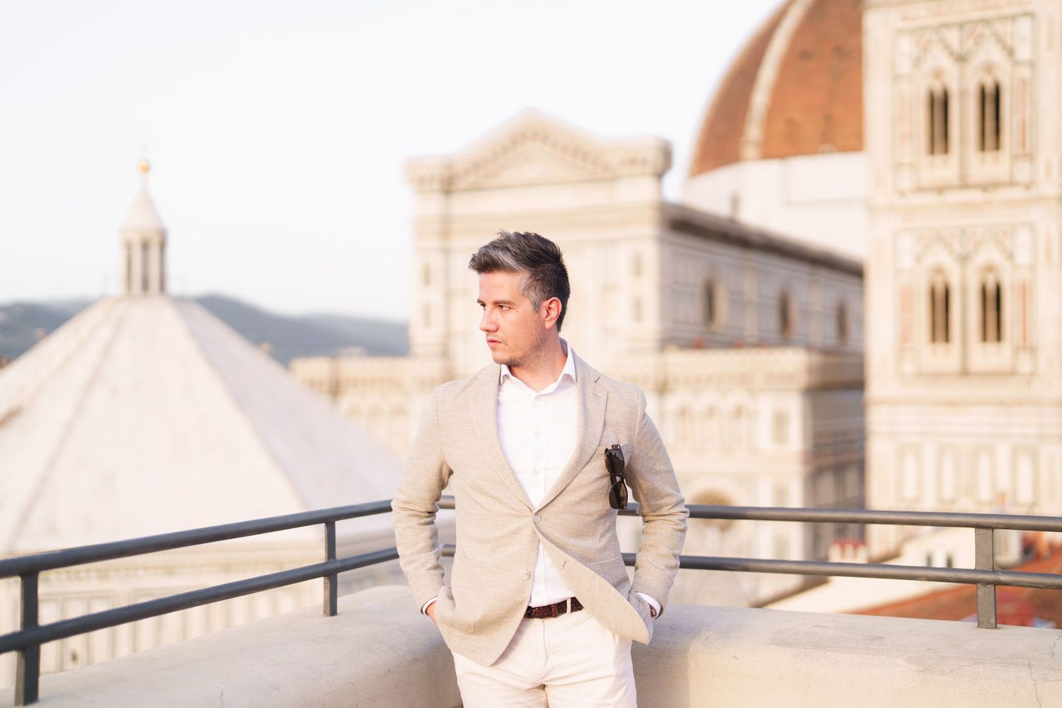 A couple in formal wedding attire embraces on a rooftop terrace overlooking a European cityscape at sunset.