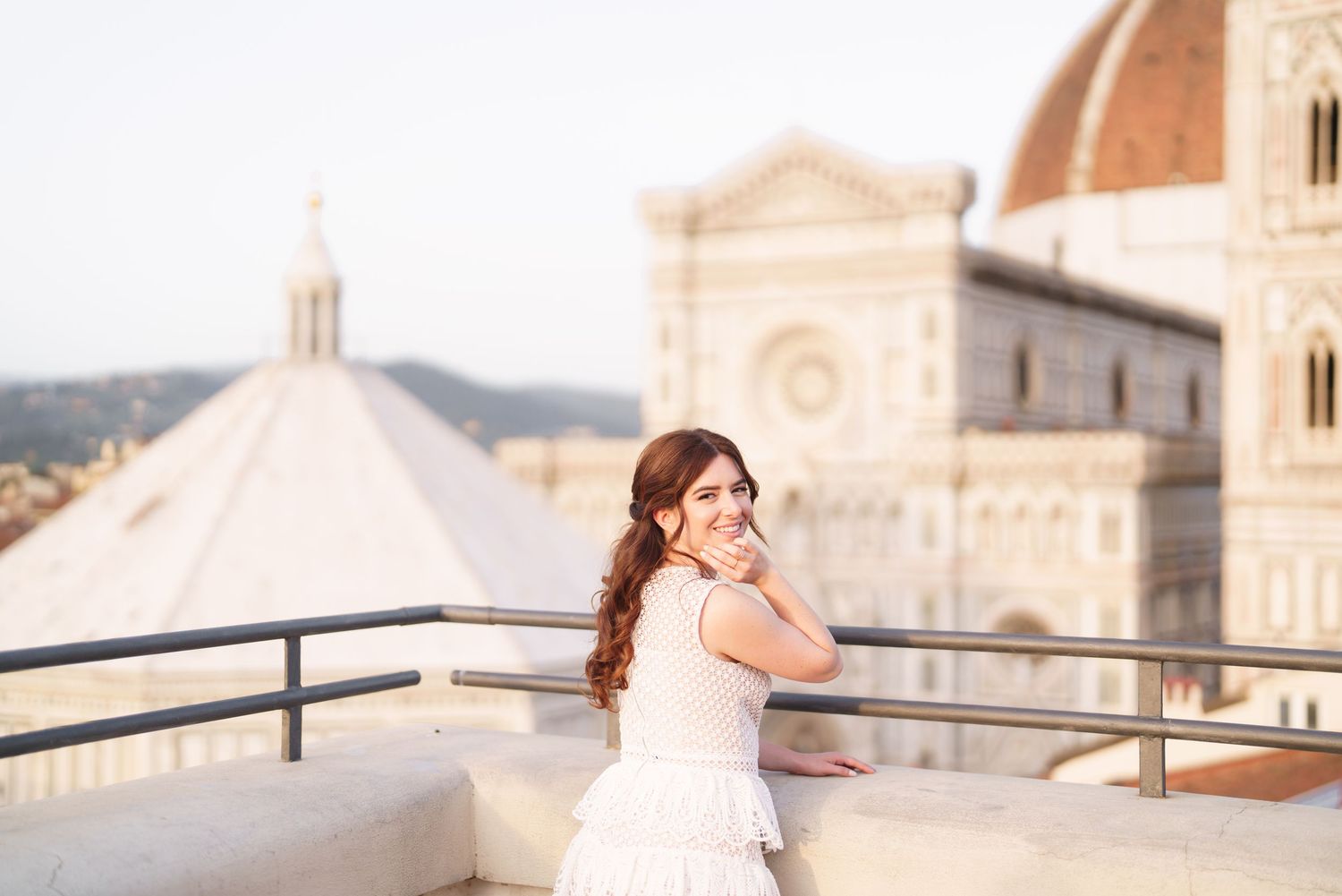 A bride in a white dress poses gracefully on a balcony overlooking a European city during golden hour.