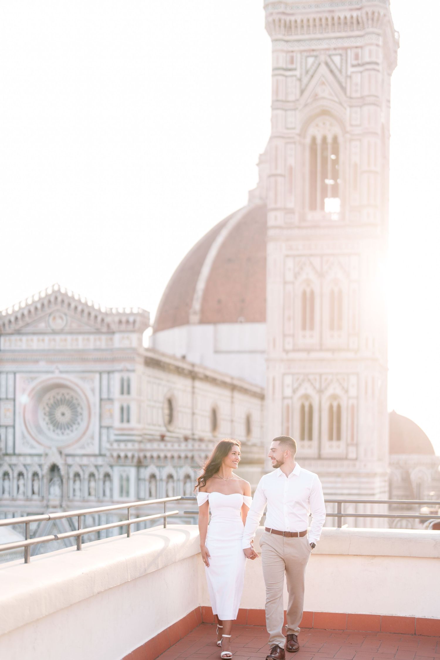 Couple in white outfits walking on rooftop terrace with Florence Cathedral's iconic dome and architecture in background.