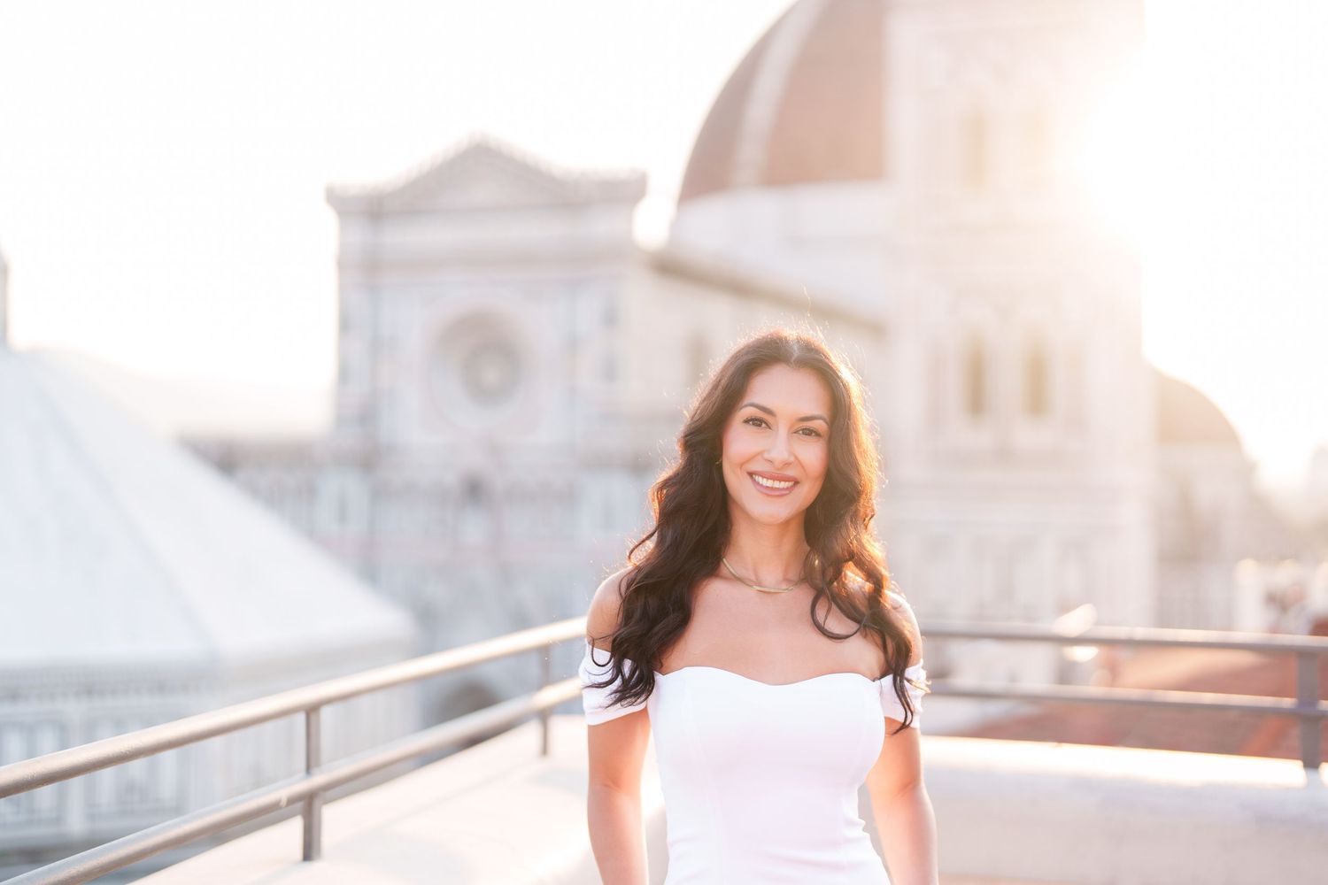 Woman in white dress poses on stone steps during golden hour sunset with architectural backdrop.