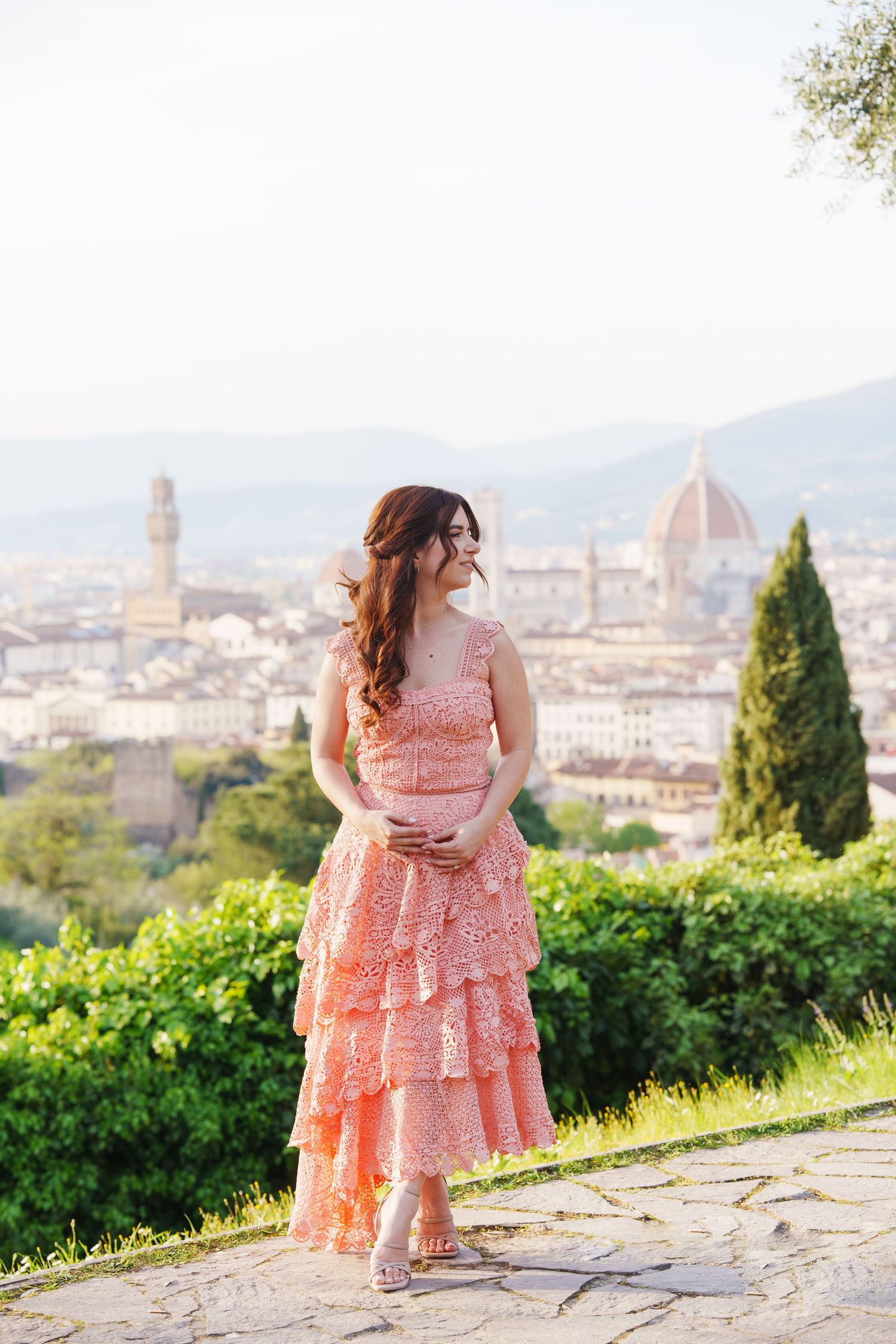 A person in a coral lace dress poses at a scenic overlook with Florence, Italy's iconic Duomo cathedral in the background.