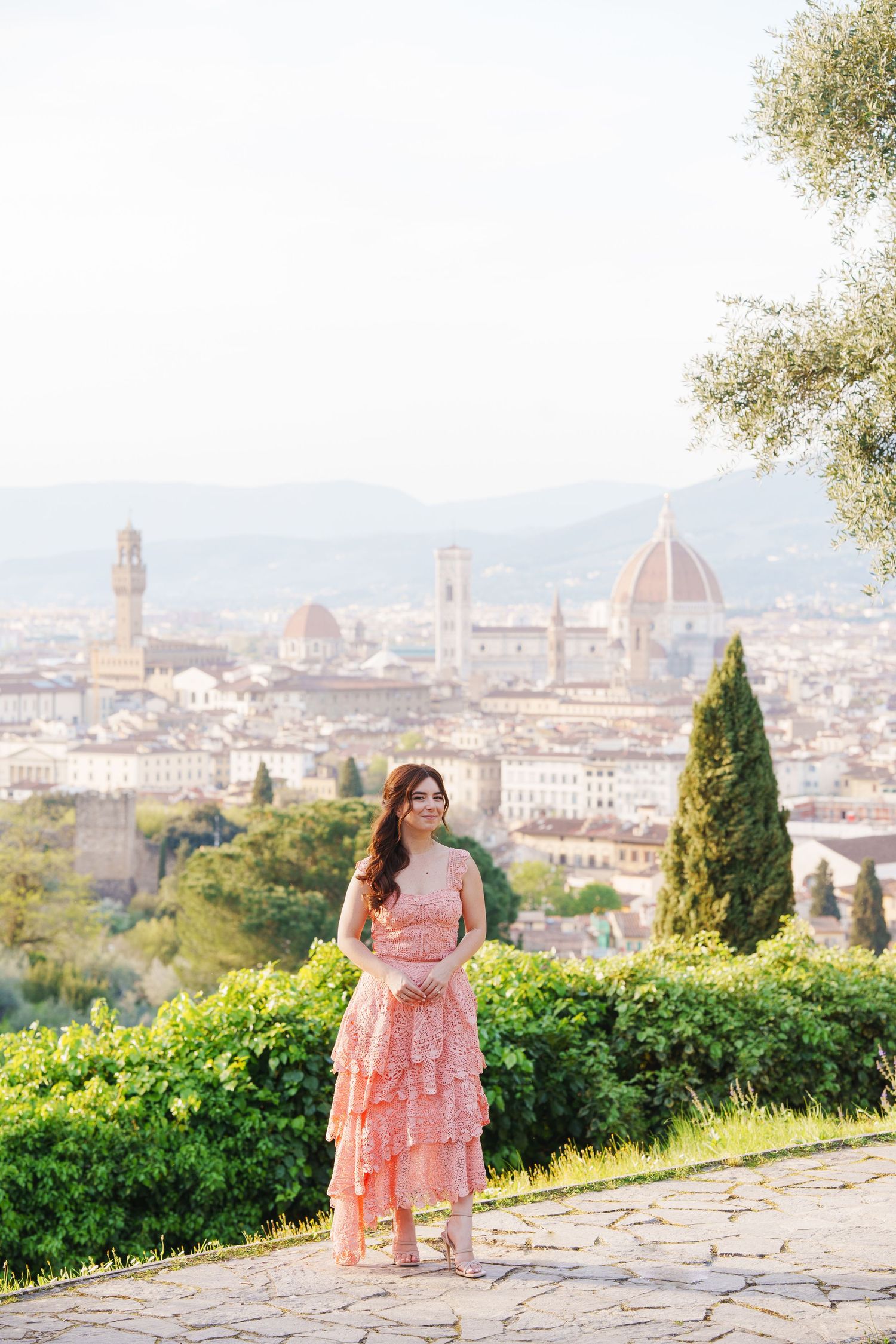 A person in a pink dress poses against the scenic backdrop of Florence, Italy with its iconic Duomo cathedral visible.