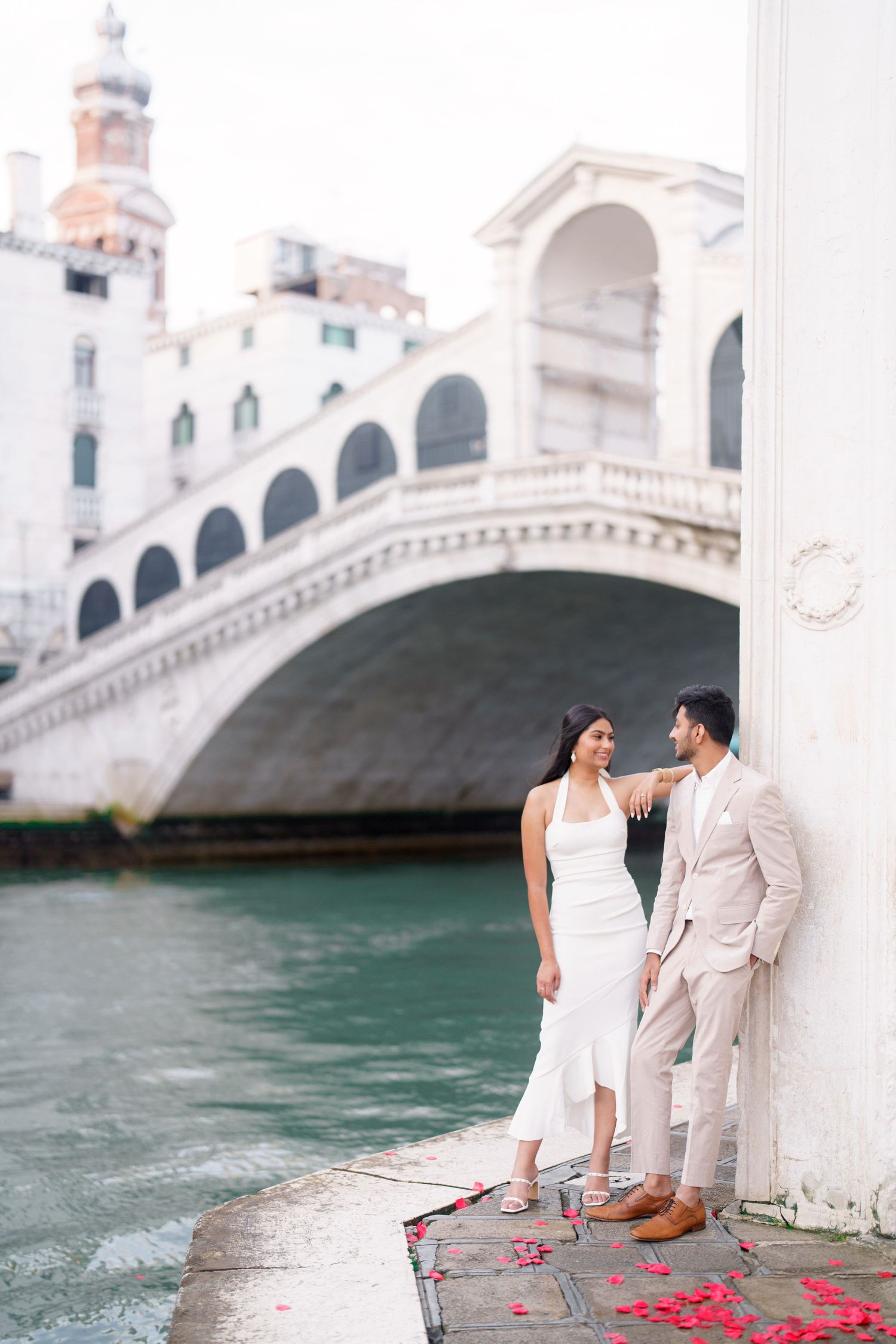 Couple sitting by Venice canal with scattered red rose petals near the iconic Rialto Bridge.