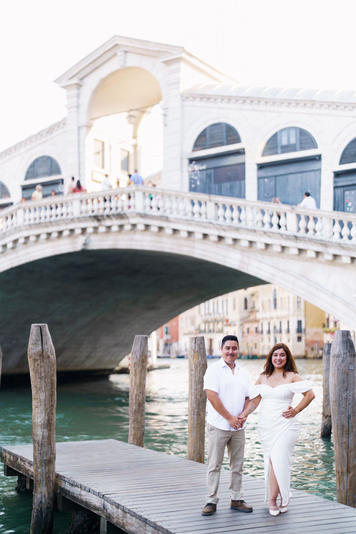A couple stands on a wooden dock near the iconic Rialto Bridge in Venice, Italy with the canal in the background.