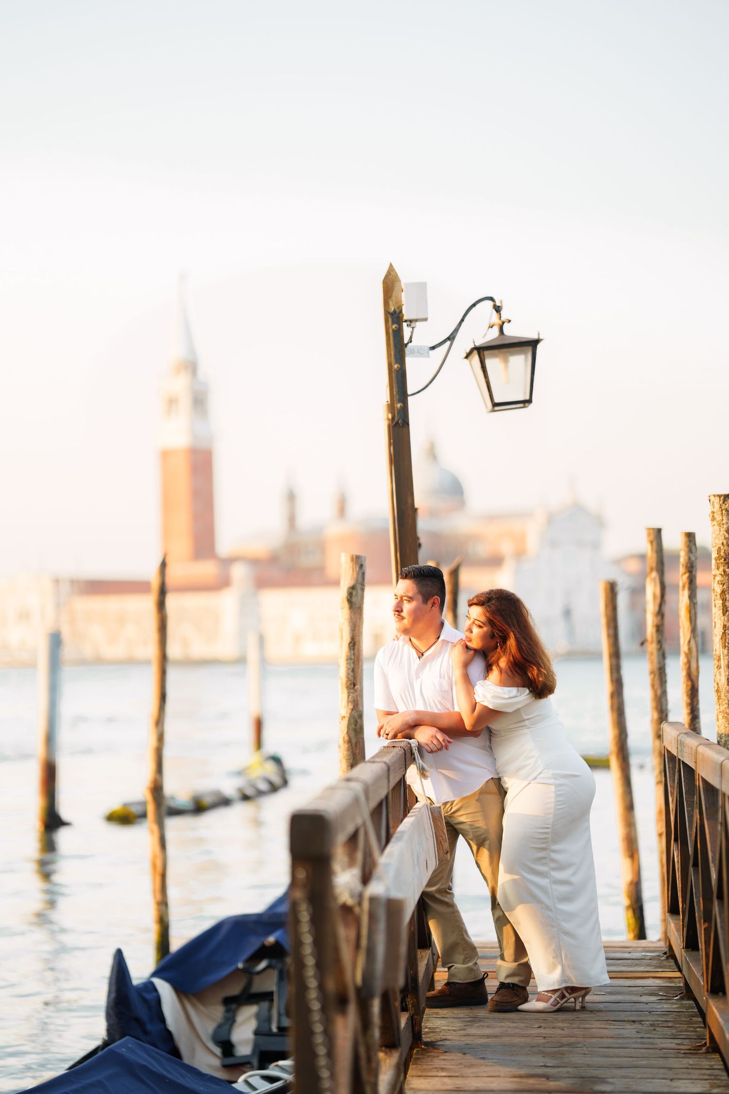photographer venice A couple embraces on a wooden pier in Venice at sunset with gondolas and San Giorgio Maggiore church in the background.