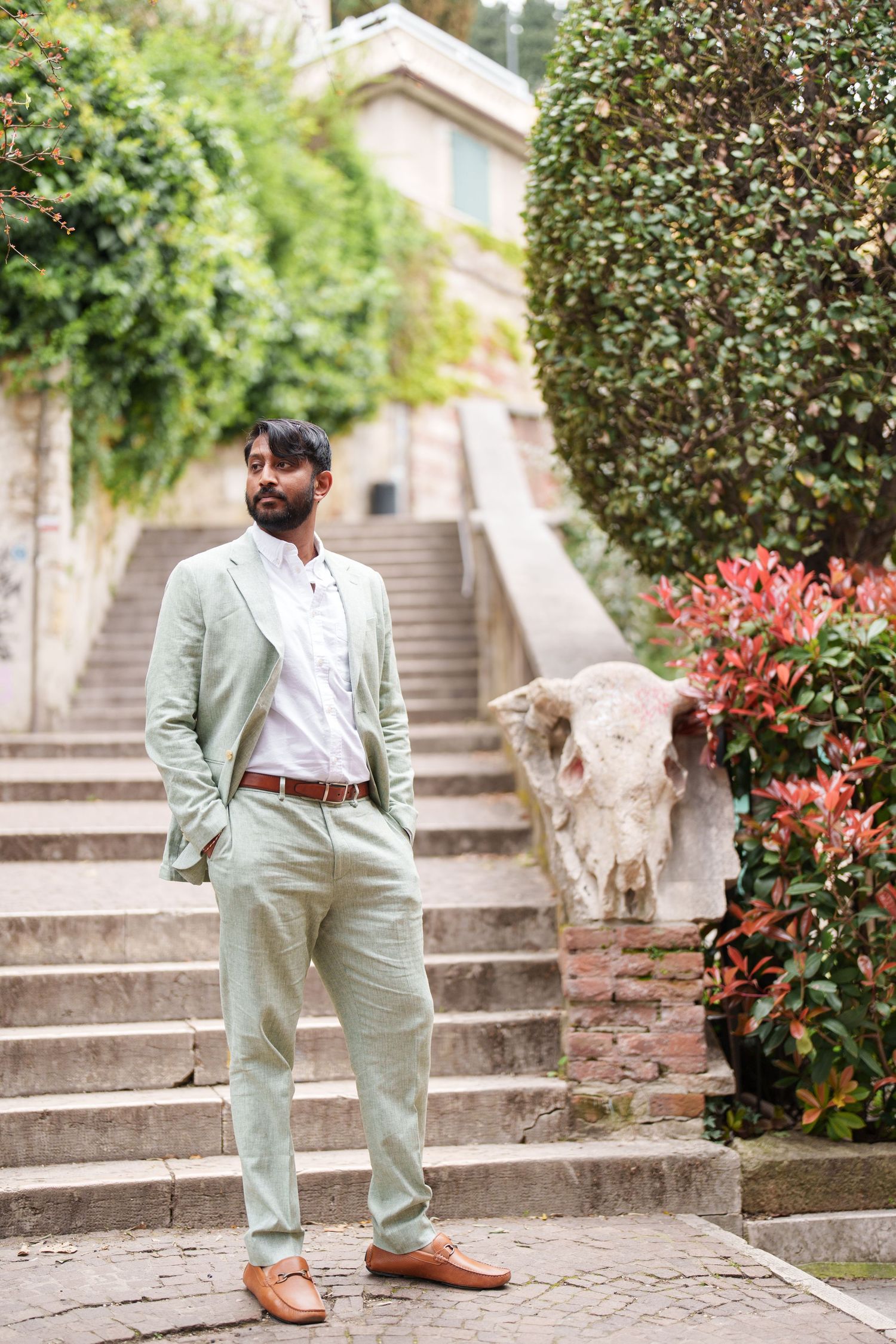 photographer verona A person in a mint green suit poses on stone steps near flowering bushes, showcasing brown leather shoes.