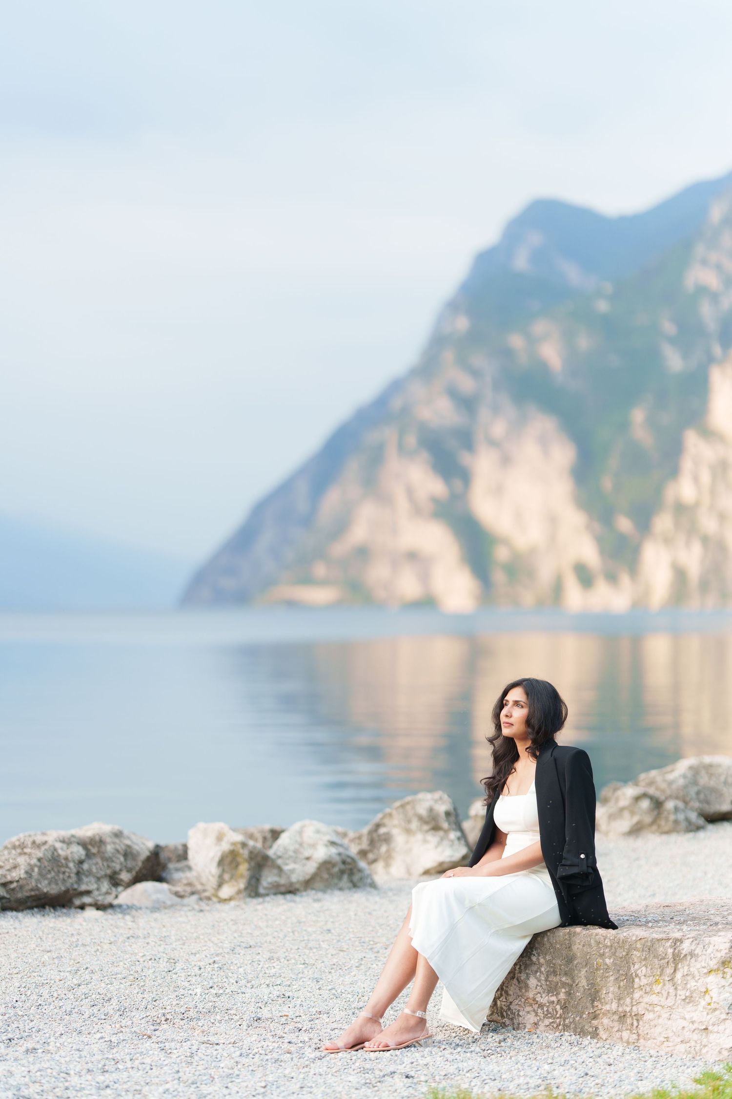 A person sits on a rocky beach shore with scenic coastal cliffs and calm blue waters in the background.