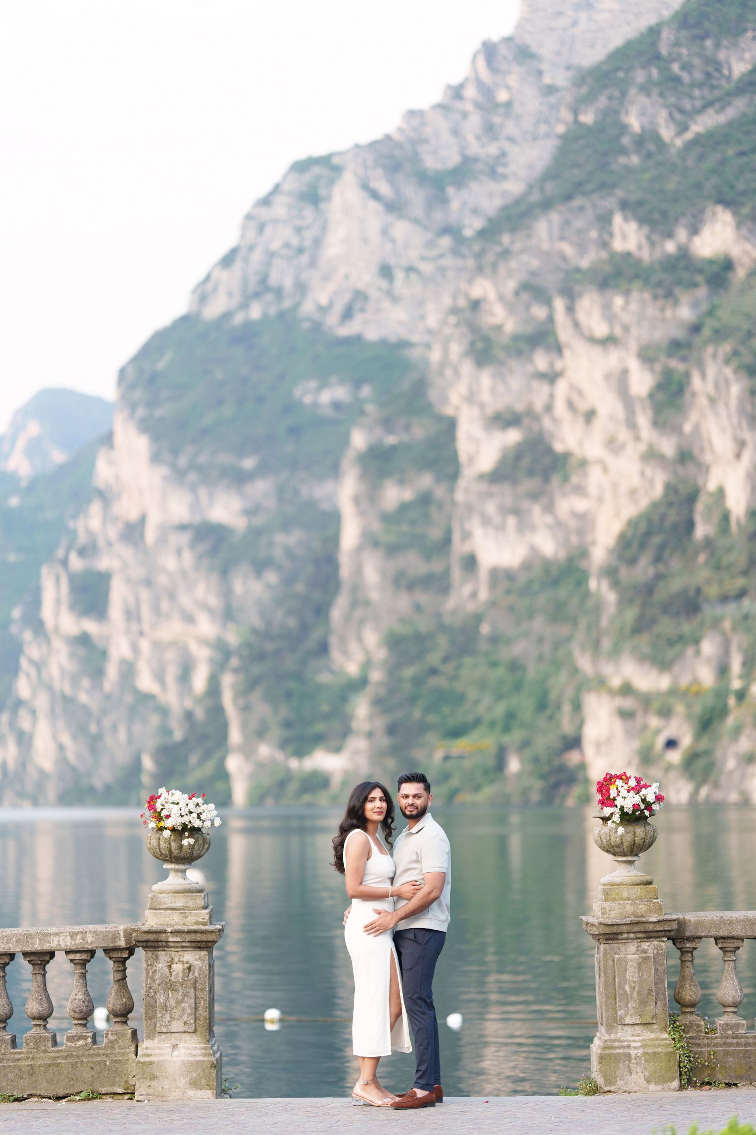 Romantic couple poses along stone balustrade overlooking scenic Lake Como with dramatic mountain cliffs in Italy.