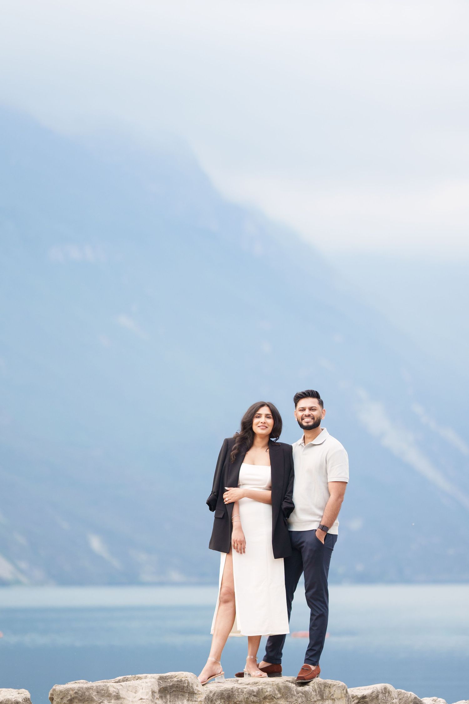 A couple stands together against a scenic mountain lake backdrop on a cloudy day.