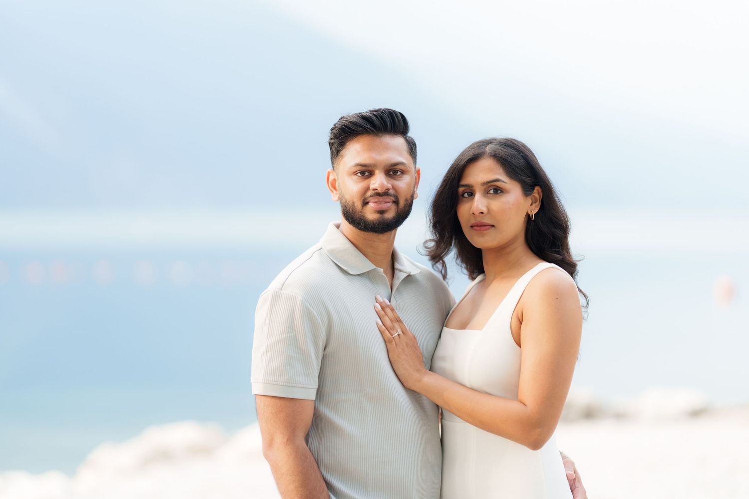 A couple poses together outdoors against a soft cloudy backdrop near the ocean.