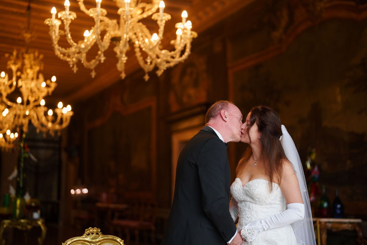 A couple shares a romantic kiss under an elegant crystal chandelier during their wedding reception.