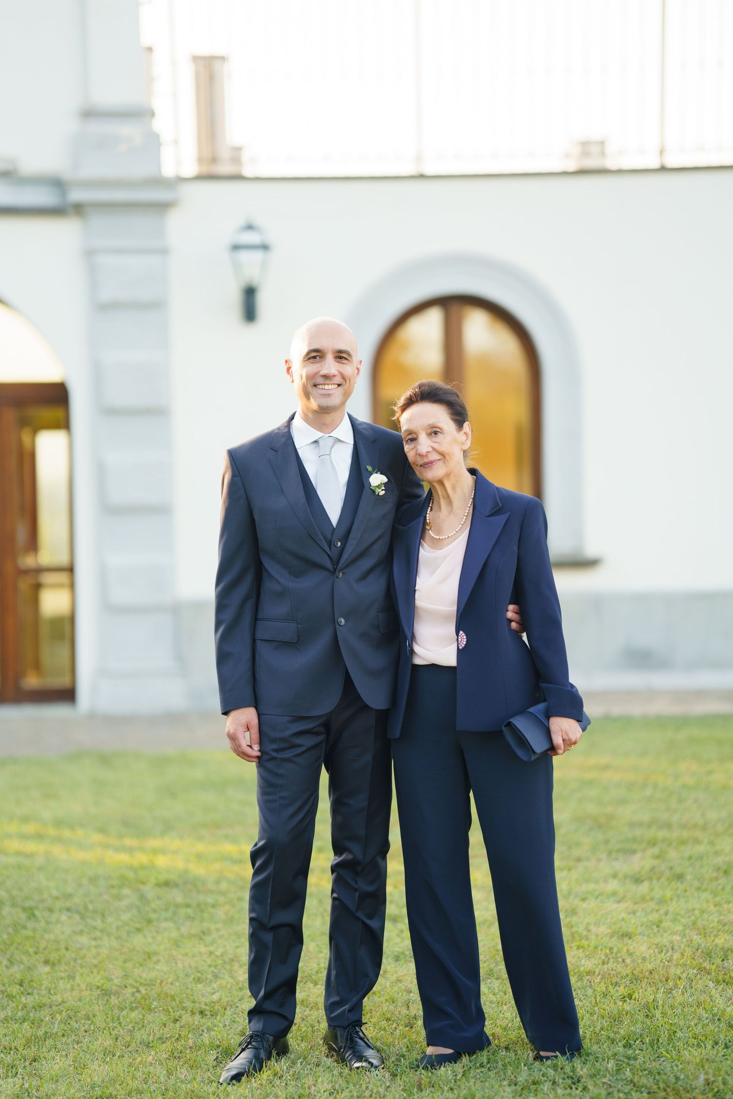 Two people in navy blue suits standing together on grass near a white building.