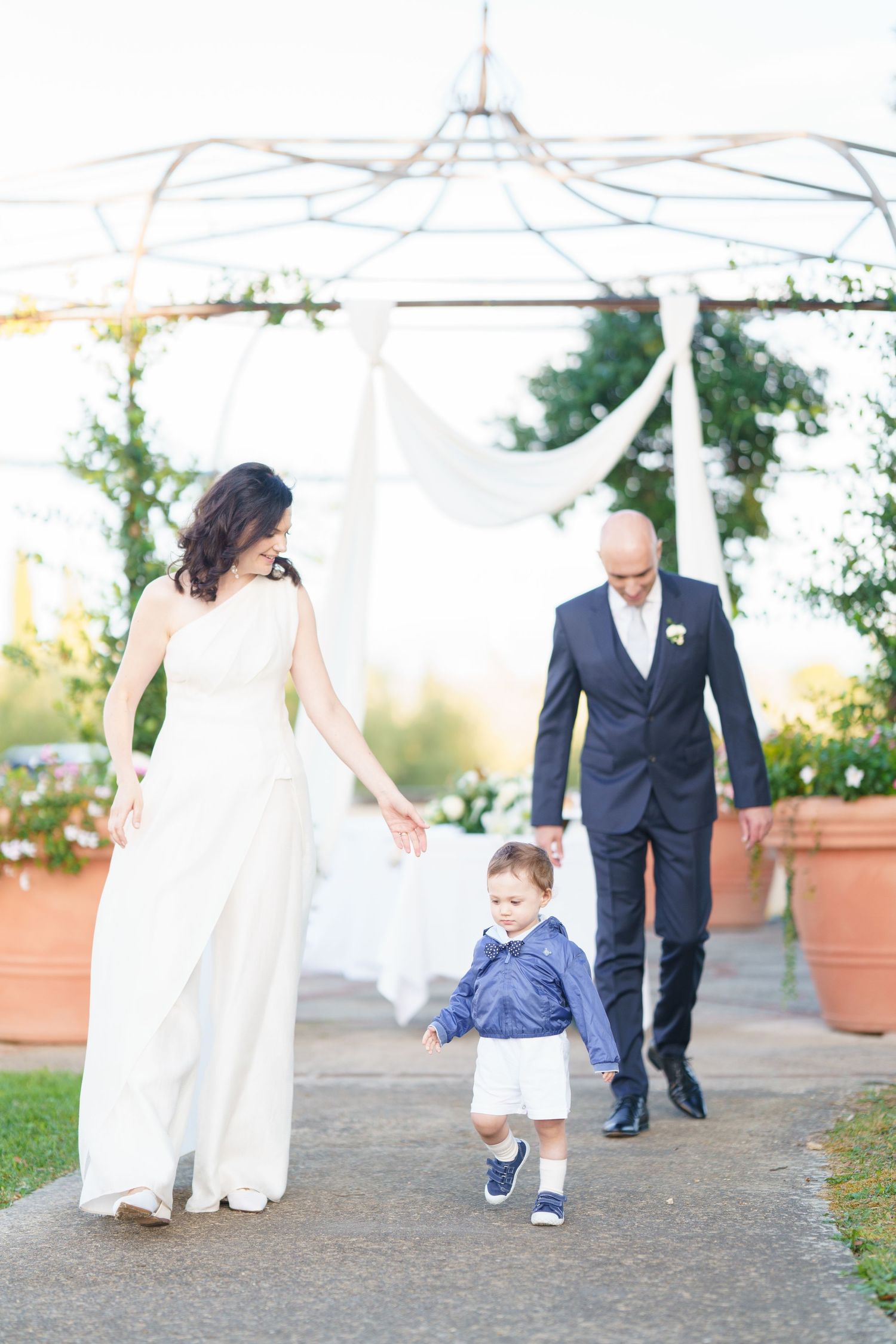 Outdoor wedding scene with couple and child walking on path under white canopy.
