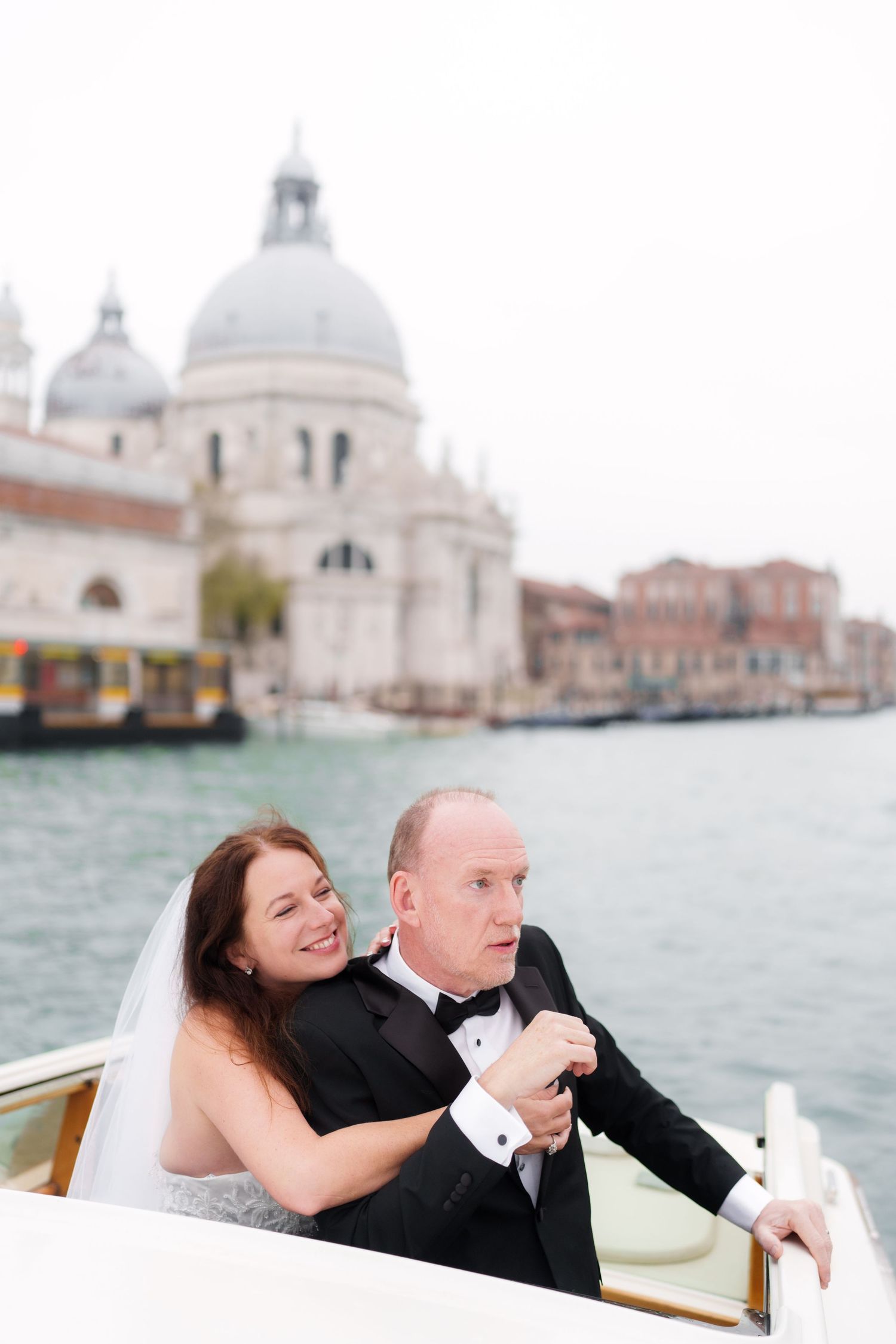 A romantic couple shares joyful moments on a water taxi in Venice with the iconic Santa Maria della Salute church in view.