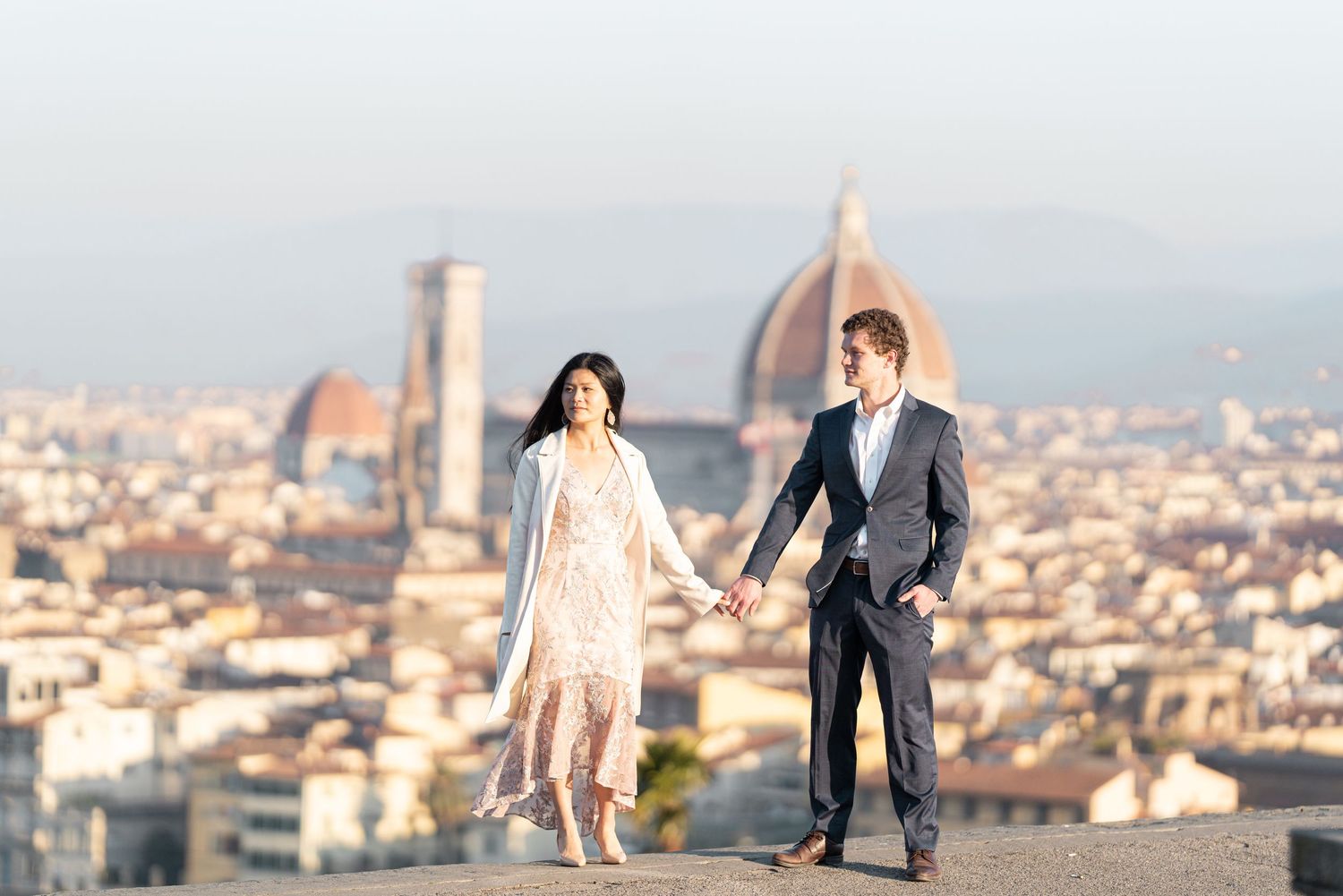 Romantic couple stands on balcony overlooking Florence's iconic Duomo cathedral and historic cityscape at sunset.